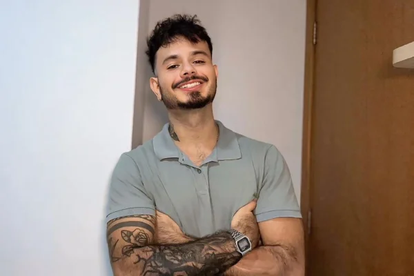 Foto colorida de homem sorrindo de braços cruzados em frente a uma parede branca - Metrópoles.