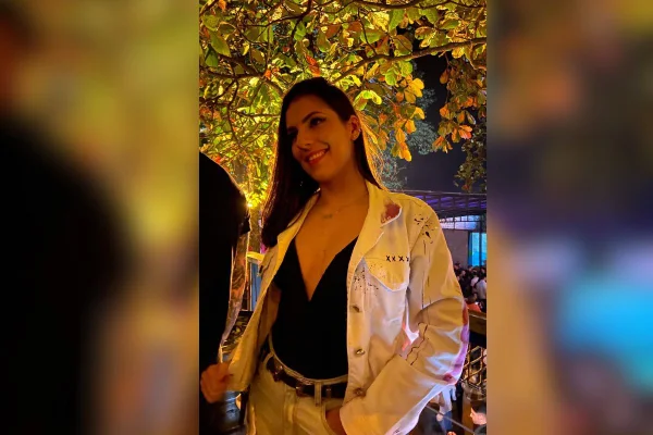 imagem colorida mostra mulher de cabelo liso e escuro sorrindo. ela é branca, veste uma regata preta, uma jaqueta branca, e uma calça branca com cinto escuro. atrás dela está uma árvore. metrópoles