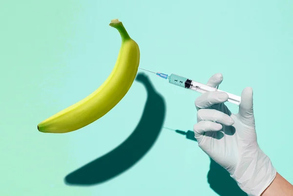 Mão do médico segurando uma seringa injetando fluido em uma banana em fundo azul claro. Conceito de disfunção erétil