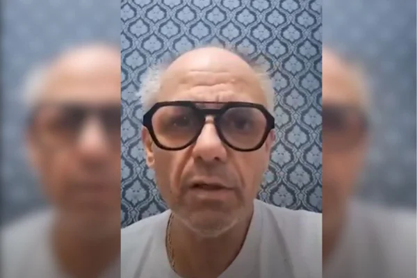 imagem colorida mostra homem branco, de óculos de grau. ele tem cabelos brancos, usa camiseta branca e está na frente de um papel de parede azul - metrópoles