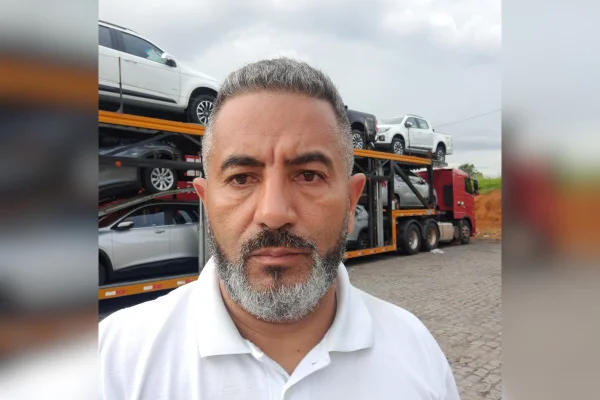 imagem colorida mostra homem com cabelo grisalho e camisa branca. ele está em frente a uma caminhão cegonha carrregado de vários carros