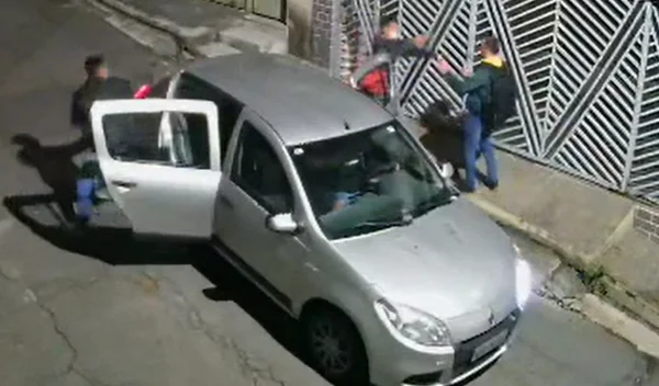 Veículo prata com portas traseiras abertas; um homem aponta arma para outro ao lado do carro - Metrópoles