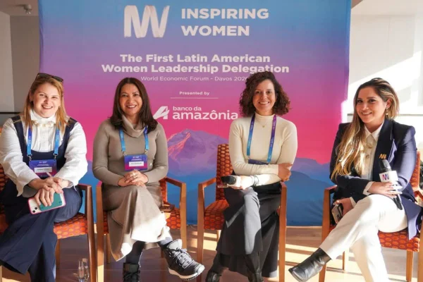 Cinco mulheres estão sentadas em cadeiras alinhadas, sorrindo para a câmera. Todas usam roupas formais e crachás. Atrás delas, um grande painel com o nome do evento reforça o contexto institucional e internacional