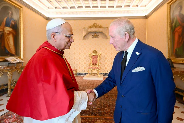 Foto colorida de homem com vestes de papa apertando a mão de homem com terno azul - os homes são o rei Charles III e papa Leão XIV - Metrópoles