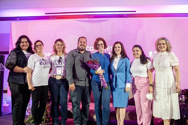 Grupo de pessoas em frente a telão com cor rosa. Uma mulher segura um buquê