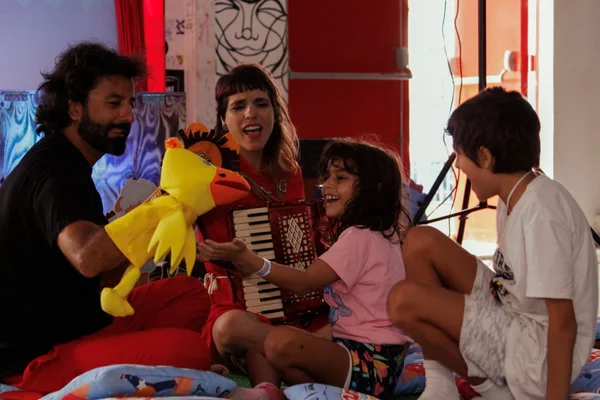 Imagem colorida mostra criança e adultos brincando com um papagaio amarelo de pelúcia - Metrópoles