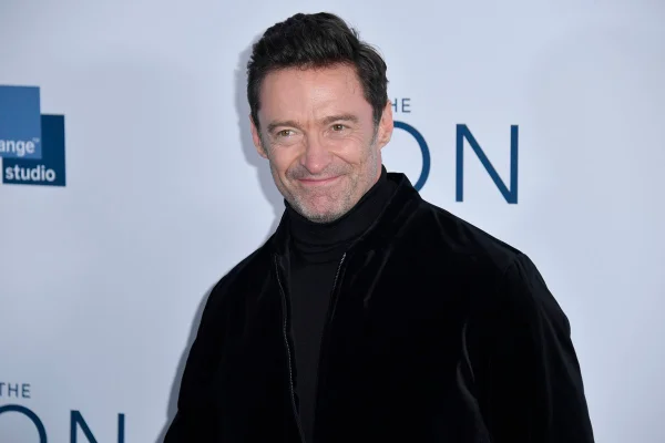 Hugh Jackman é um homem branco, de 55 anos, alto, cabelos pretos. Ele está usando uma roupa toda preta e dá um leve sorriso para a câmara - Metrópoles