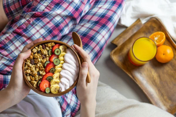 Imagem colorida de pessoa sentada na cama, comendo frutas