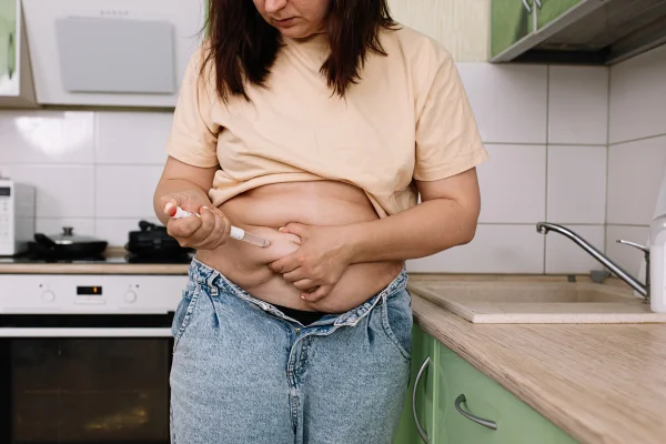 Foto colorida de mulher em cozinha aplicando medicamento na barriga - Metrópoles