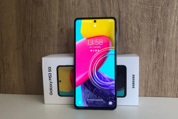 O celular Samsung Galaxy M53 5G com a tela de bloqueio ligada, em pé e encostado em sua embalagem - Metrópoles