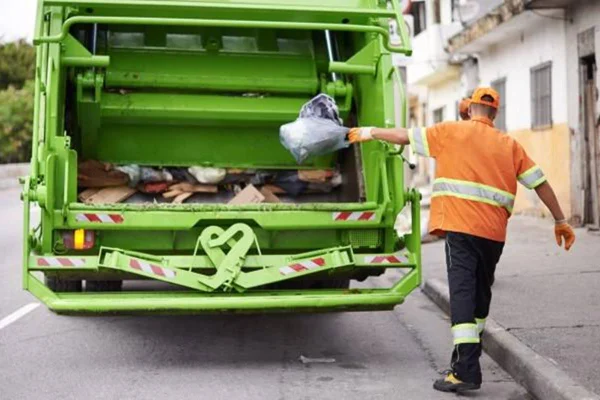 Foto colorida de homem com colete laranja jogando lixo em traseira de caminhão de lixo