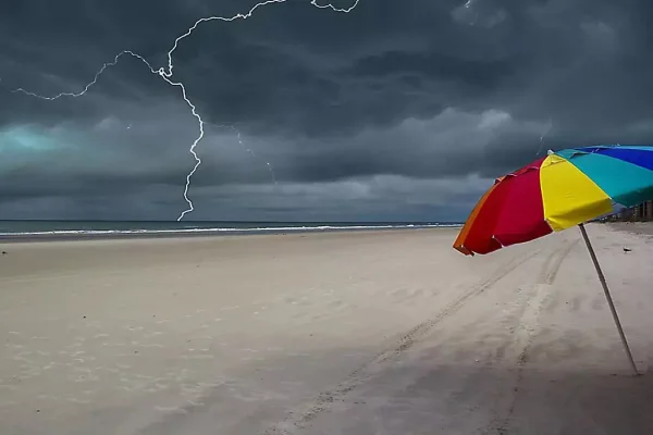 Imagem colorida de um raio em uma praia, que tem um guarda-sol colorido fincado na areia