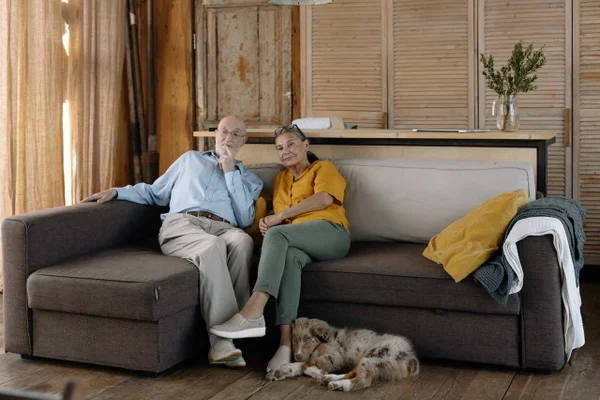Homem e mulher idosos, sentados em um sofá escuro juntos, com um cachorro deitado próximo aos pés.