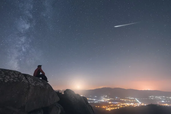 foto colorida de céu azul escuro, um homem ao lado esquerdo sentado em uma montanha observando uma pequena cidade de longe - Metrópoles.