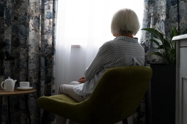 Foto colorida de mulher idosa, sentada em um quarto sozinha, enquanto olha pela janela - Metrópoles.