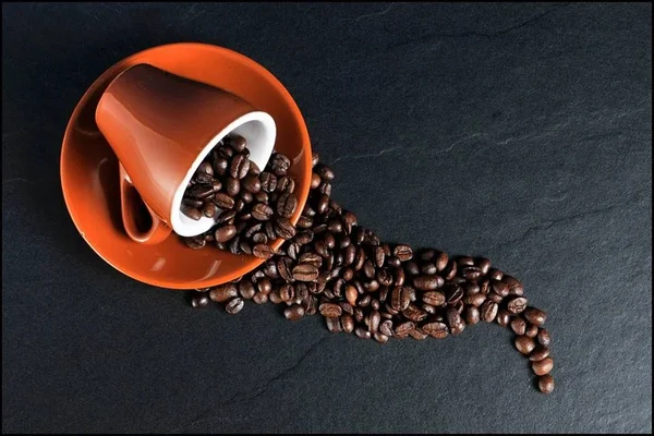 Caneca laranja dentro de tigela da mesma cor com grãos de café saindo sob fundo preto - Metrópoles