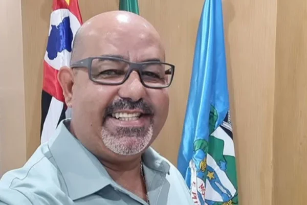 Imagens coloridas mostram homem sorrido, careca raspada, óculos, posando à frente das bandeiras de São Paulo, do Brasil e de Mongaguá