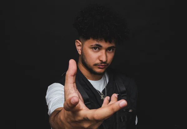 DJ Klap. Ele é um homem branco, com cabelo e barba preto. Usa um colete preto, uma camisa branca e está com a mão para a frente. O fundo é preto - Metrópoles