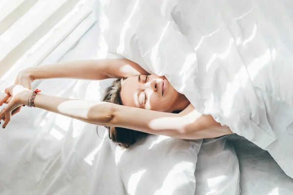 Na cama, embaixo de lençol banco, mulher acorda e se espreguiça - Metrópoles