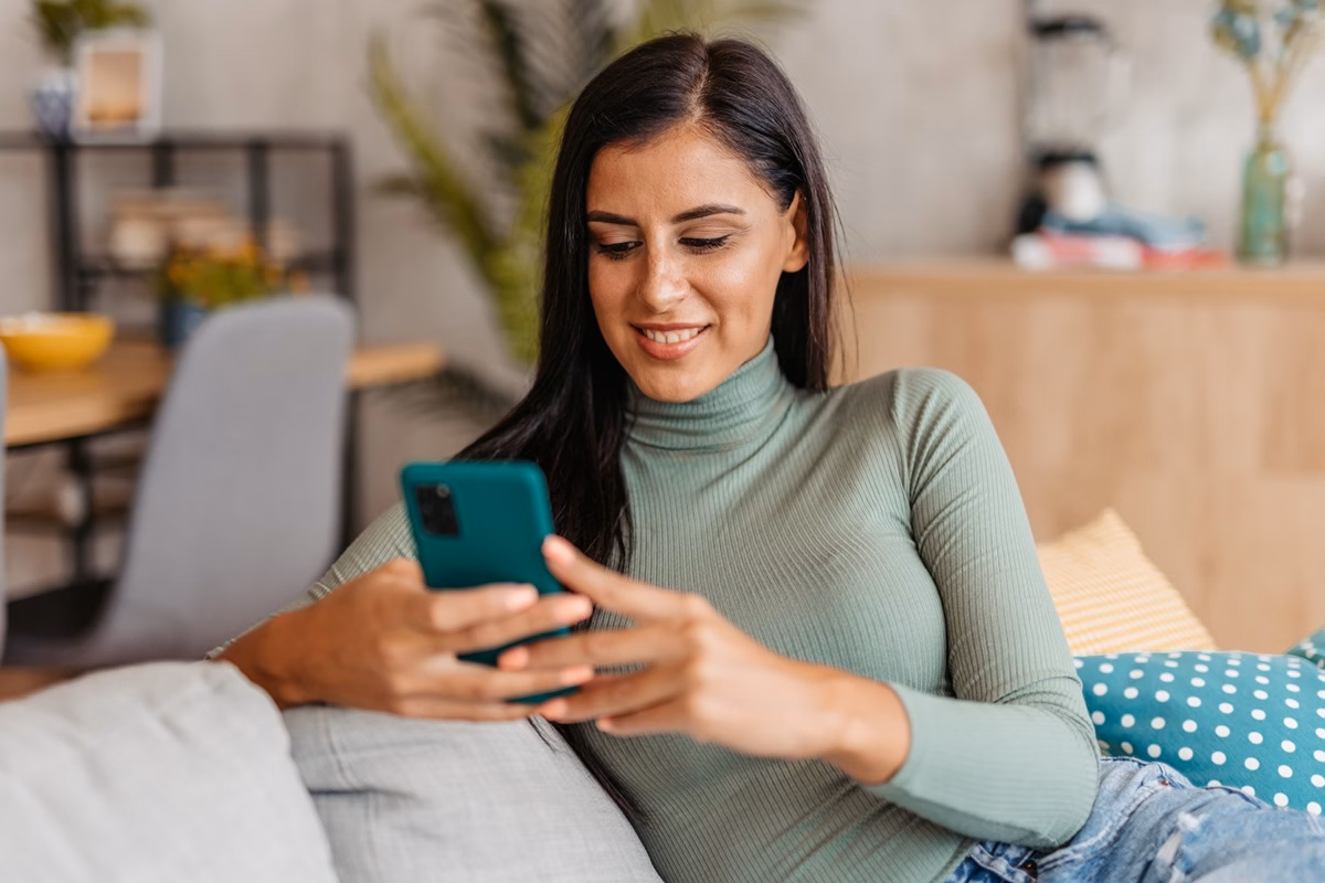 Sorridente jovem relaxando em casa, enviando mensagens de texto no smartphone.