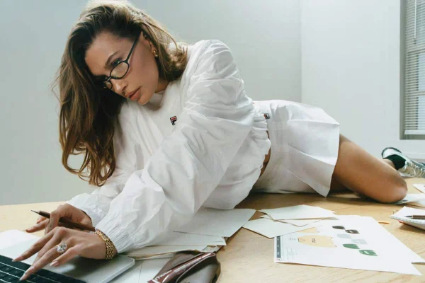 A imagem mostra a modelo Hailey Bieber sob uma mesa de escritório com papei espalhados