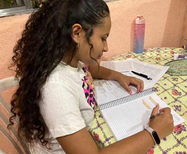 Imagem colorida mostra Deborah Maria da Silva Santos, uma mulher de pele morena, cabelo comprido cacheado preto, usando uma camiseta branca, segurando uma caneta e olhando para um caderno. Ela é uma das estudantes que vai fazer o Enem em 2023 - Metrópoles