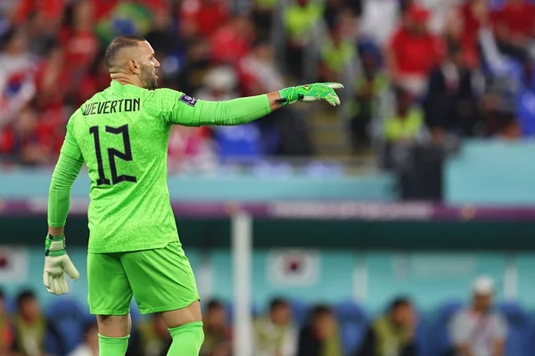 Homem branco e com uniforme de goleiro do Brasil. De costas para a foto, ele está em pé e apontando para algo dentro de campo durante partida de futebol na Copa do Mundo. Ao fundo, o estádio de futebol cheio e com pessoas assistindo ao jogo em desfoque