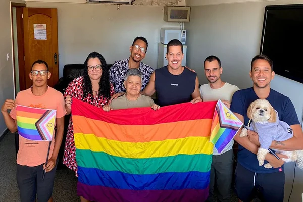 Sete pessoas e estão segurando uma bandeira LGBT. A foto ainda tem um cachorro no colo de um homem à direta