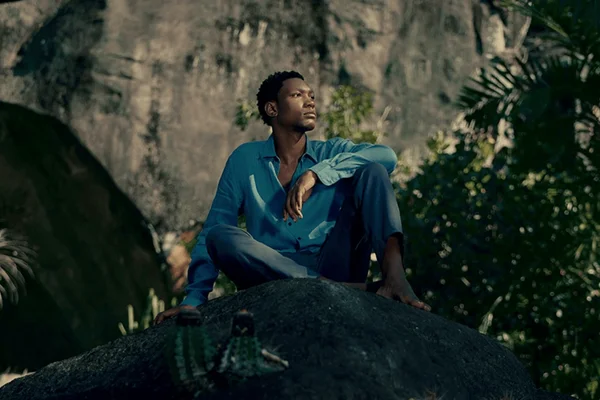 foto com cor. modelo negro sentado em pedra em meio a natureza com roupa de alfaiataria - metrópoles