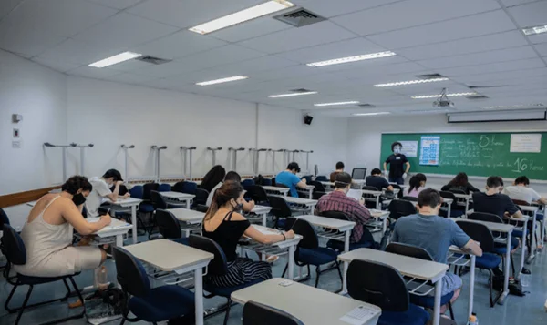 Imagem colorida, pessoas sentadas em carteiras realizando uma prova