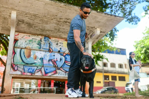 Foto colorida de homem negro na parada de ônibus puxando um cão-guia da raça labrador e da cor preta