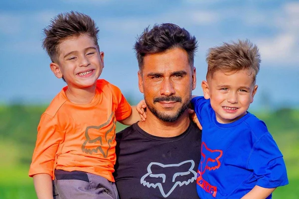 Julio Rocha e os filhos, José e Eduardo. Ator usa roupa preta e segura os meninos, que usam laranja e azul, no colo - Metrópoles