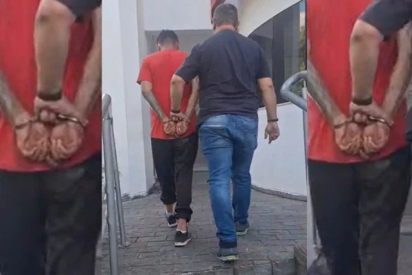 Homem de camiseta vermelha, algemado com as mãos para trás, é conduzido por policial, estando ambos de costas no registro - Metrópoles
