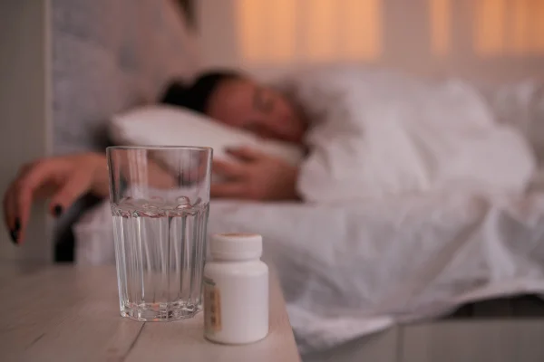 Foto colorida de mulher deitada na cama com remédios para dormir em cima da mesa - Como os remédios para dormir agem no corpo