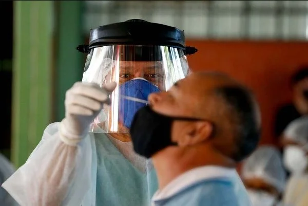Na imagem colorida, dois homens estão posicionados no centro. Um está sentado enquanto outro está em pé, com máscara, roupa de hospital na cor azul e coloca algo semelhante a um cotonete no nariz do moço sentado