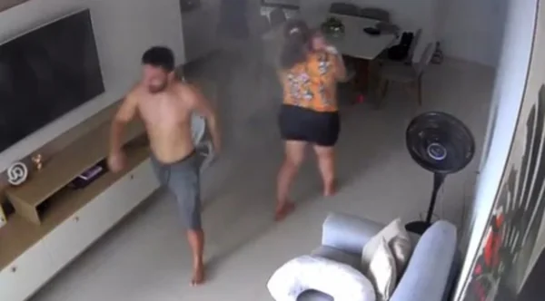 Mulher com criança no colo e homem de bermuda em sala do apartamento