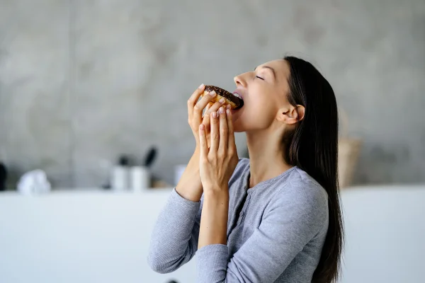 Retrato de uma mulher alegre saboreando um donut doce delicioso em casa. Conceito de comida não saudável. Metrópoles