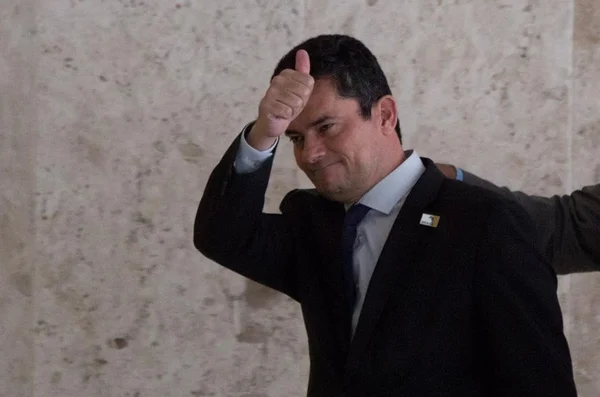 Na imagem colorida, um homem está posicionado à esquerda. Ele veste blazer escuro e gravata preta. Ele possui cabelo preto