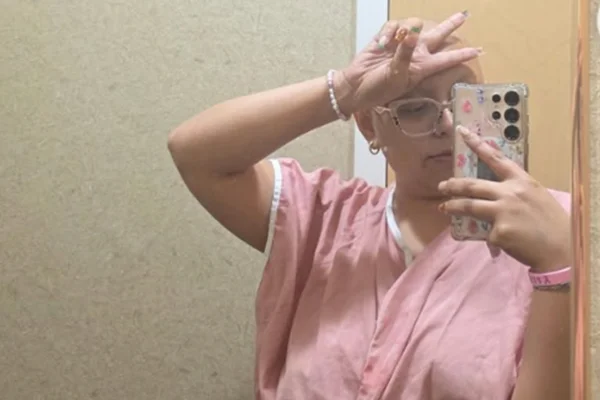 Foto colorida de mulher com avental rosa, fazendo sinal com os dedos, enquanto tira foto do celular pelo espelho - Metrópoles.