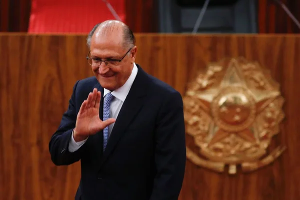 Na imagem colorida, um homem está posicionado no centro. Ele usa blazer escuro, camiseta branca, óculos e está com a palma da mão para cima