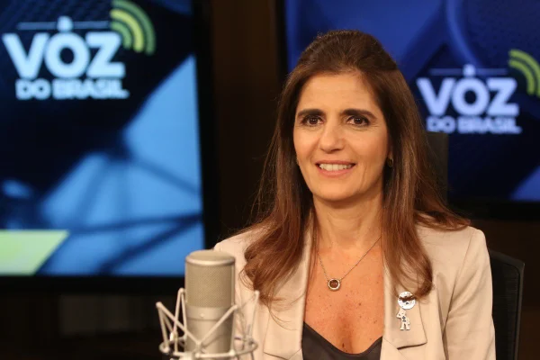 Imagem colorida de Ana Estela Haddad, mulher branca, de cabelos castanhos na atura do ombro, de blazer beje e blusa decotada marrom. Ela está de frente para um microfone, com TVs ao fundo, no que parece ser um estúdio de um programa de entrevistas - Metrópoles
