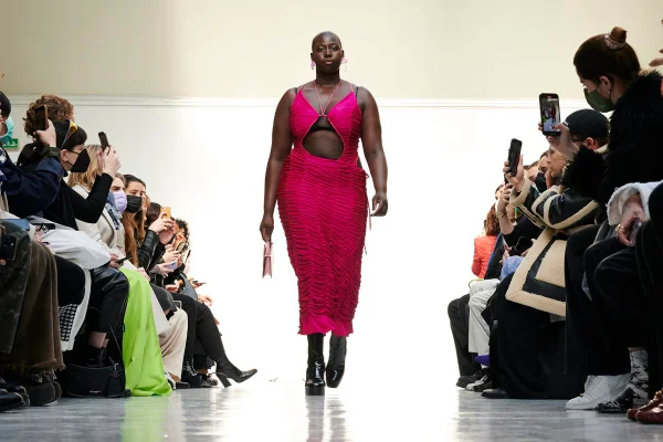 Mulher negra e careca desfilando. Ela usa um vestido franzido rosa da marca Ester Manas