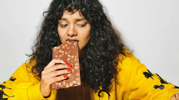Foto colorida de mulher com cara de desconfiada comendo uma barra de chocolate (compulsão alimentar)