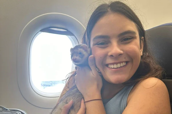 Mulher branca com cabelos escuros lisos segura furão pequeno colado ao rosto. Eles estão dentro de avião, em frente à janela