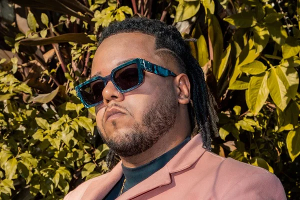 Foto colorida do rapper Vulgo FK. Ele está de óculos escuros e usa um terno rosa, com blusa preta por dentro. Plantas ao fundo - Metrópoles