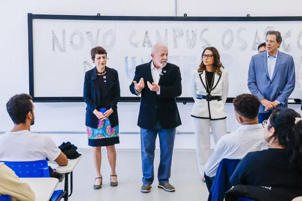 Homem branco de cabelos branco, blazer preto e calça jeans gesticula em sala de aula; ao fundo está o quadro, ao lado há outras pessoas e pé e, na frente dele, estudantes sentados em carteiras - Metrópoles