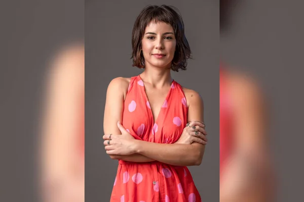Na imagem colorida, a atriz Andréia Horta aparece centralizada. Ela utiliza um vestido salmão com bolas rosas, A atriz está com os braços cruzados, possui cabelo curto e na cor escura. Ela sorri timidamente para a câmera