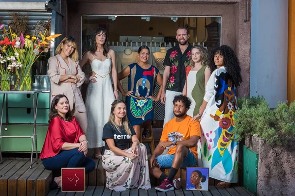 Homens e mulheres artistas organizados em uma fila para tirar foto. Cada um segura um trabalho seu, como quadros e roupas