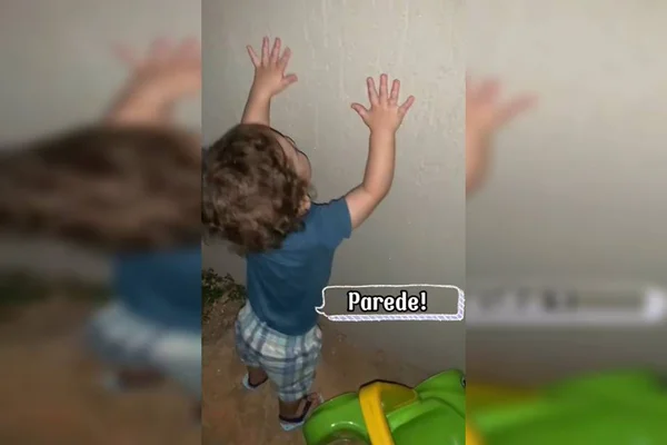 Bebê de 1,5 ano toca a parede. Ele está de roupa azul e calça xadrez também em tons azuis. Há legenda na foto com a palavra "parede"