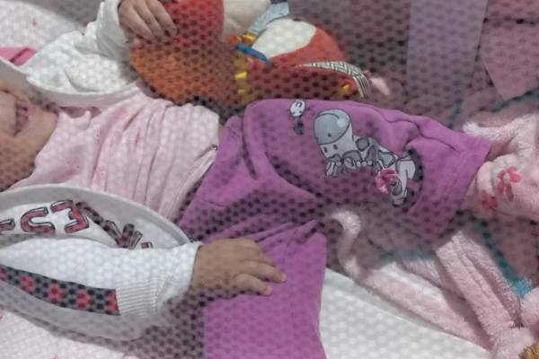 imagem colorida mostra criança em berço. a menina usa uma calça e uma blusa rosas, um casaco branco, e segura um brinquedo colorida